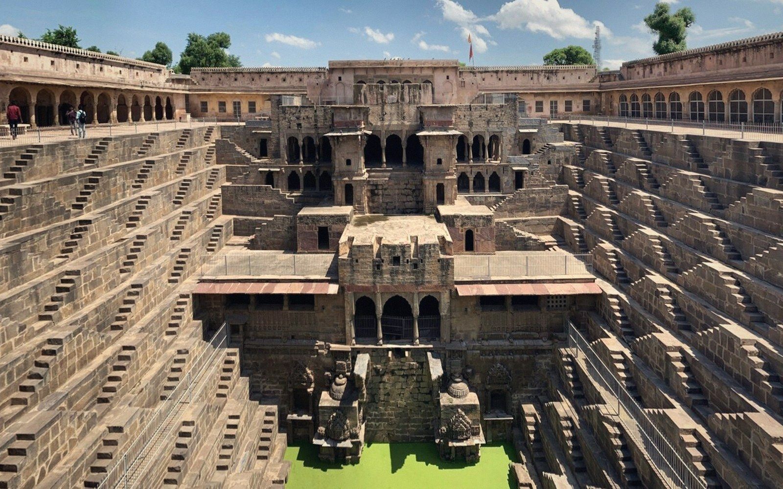 Symmetrical stepped stairways descend on multiple sides toward a green water pool at the base